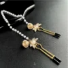 "Stellar Nipple Heart Bell Noose: A pair of heart-shaped nooses with bells and a pearl-inspired connecting chain, available in gold and silver."
